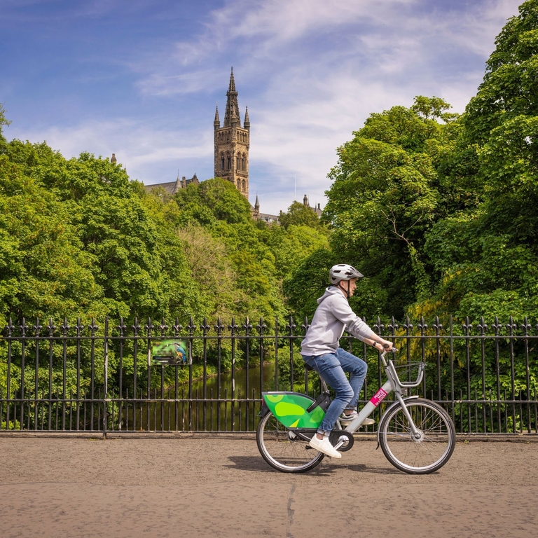 A Young Person Cycling An Ovo E-bike Over The River Kelvin in Glasgow’s popular Westend with a university and park in the background.