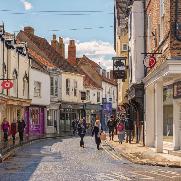 Shopping street just after a rain shower. Historic buildings line the street and shoppers walk between them. A sky with cloud is overhead.