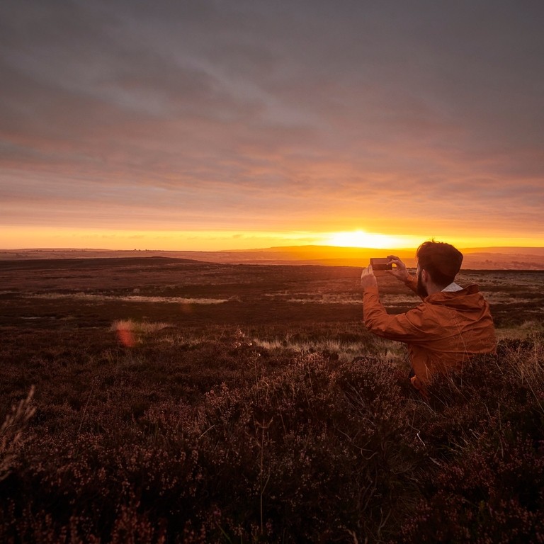 A man, wearing a red jacket, taking a picture of the sunset