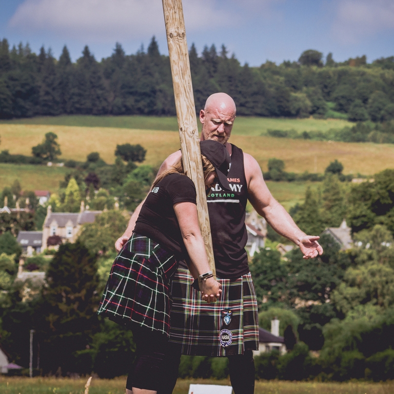 Woman wearing a kilt learning how to toss the caber with male instructor