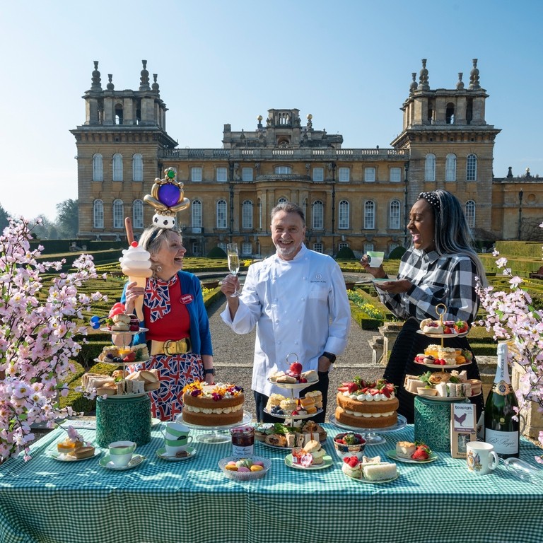 Chef and two guests stood at a display table in front of Blenheim Palace, with food and cakes on the table in front of them