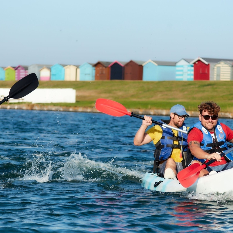 Friends kayaking on Mersea Island with beach huts in background