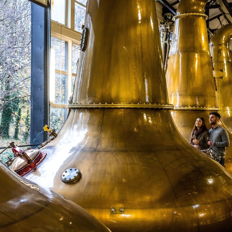 A couple standing next to the copper stills. Large windows