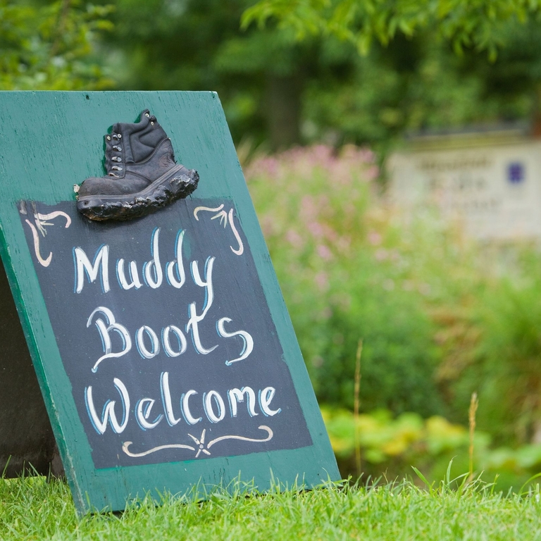 Signage on grass saying 'Muddy Boots Welcome' with plants