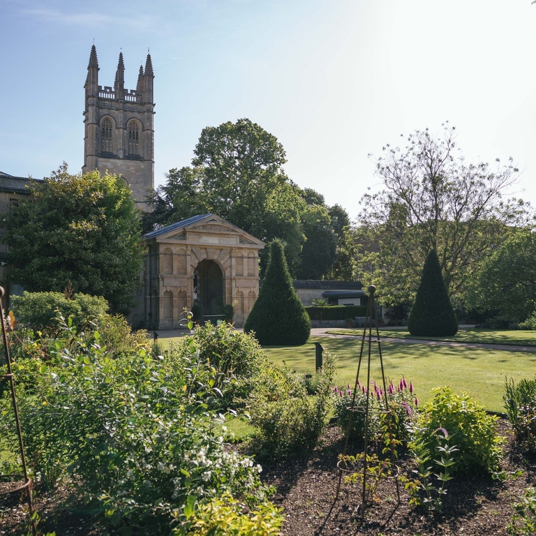 Flower bed in front of a georgian arch and building of Oxford Botanic Garden with church tower behind against blue sky