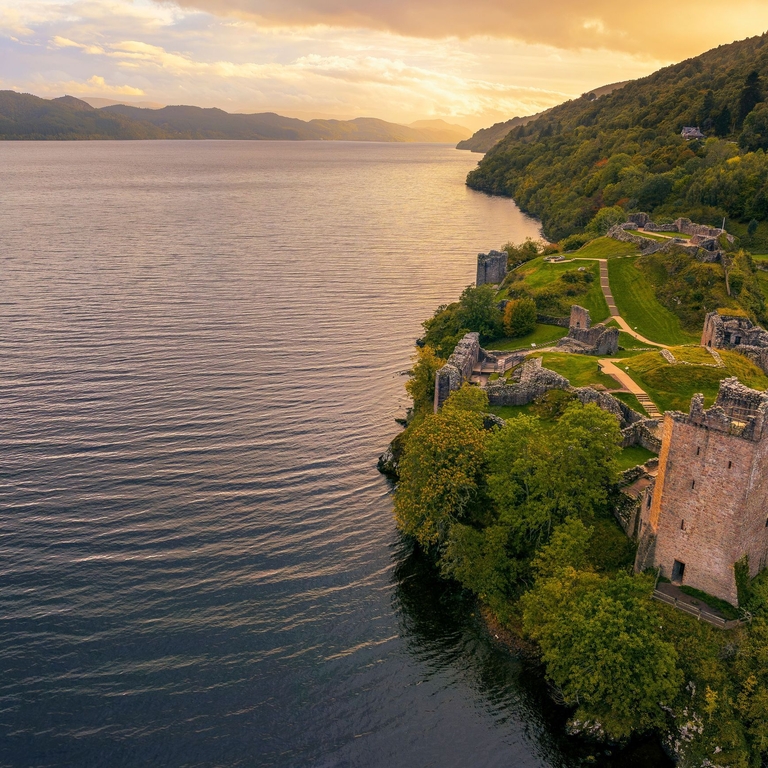 A ruined castle surrounded by trees at the edge of a lake at sunset.