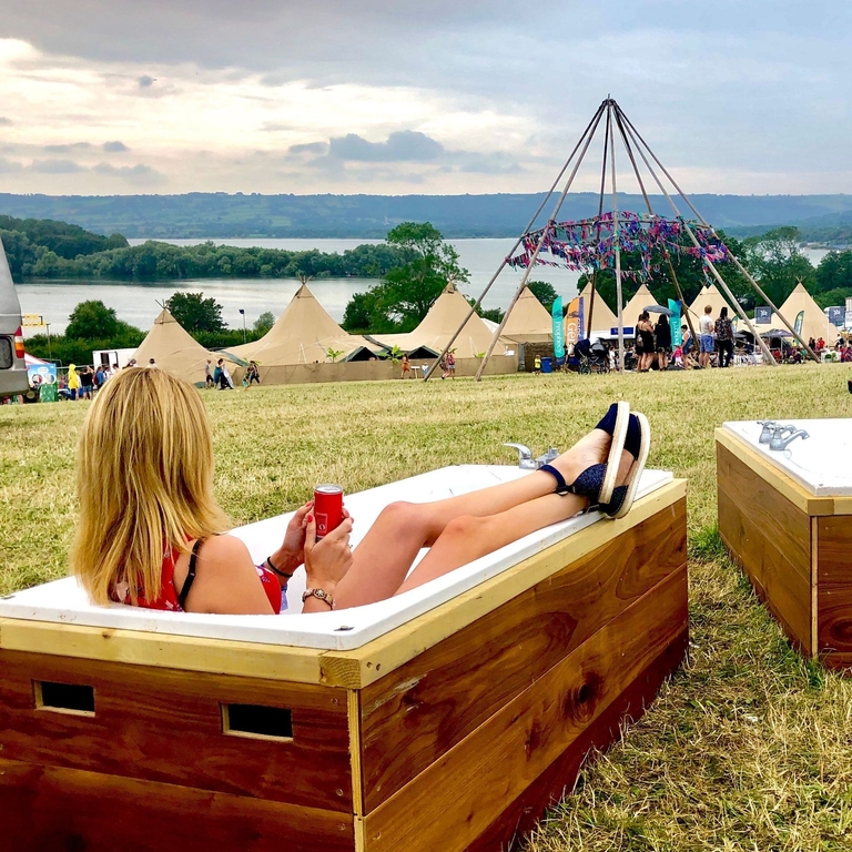 Young blond woman lying in a bath overlooking festival tents, with hills and water in the distance