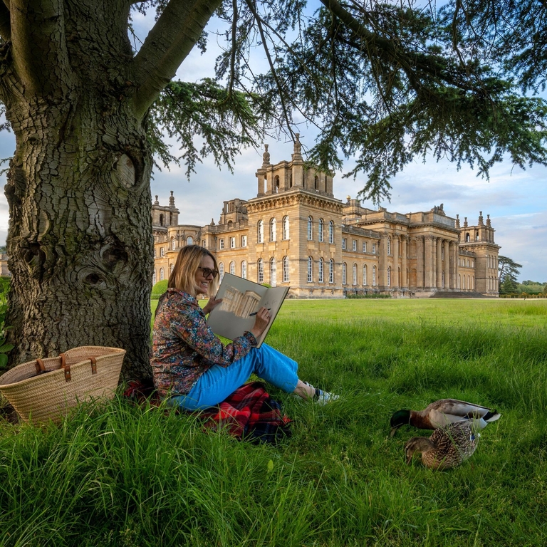 Woman sat under a tree sketching in the grounds of a large country estate