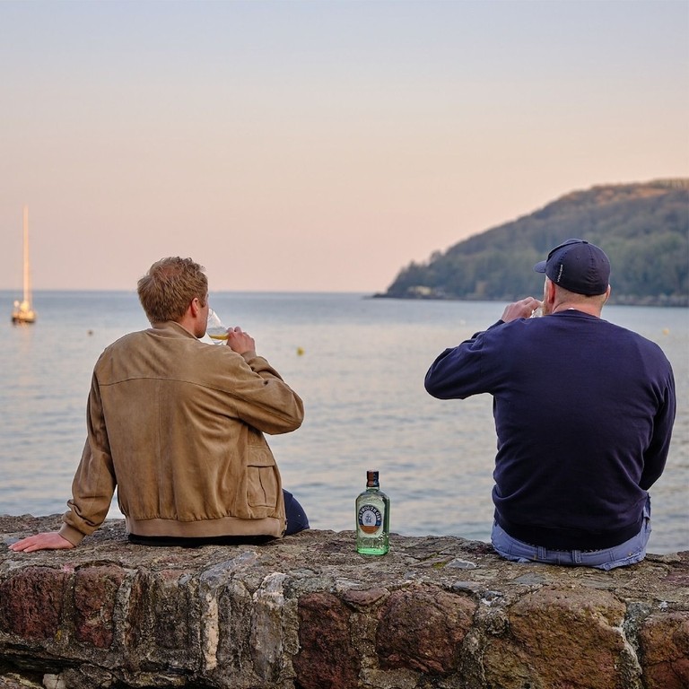 Due uomini seduti sulla parete del mare che guardano il mare bevendo Plymouth Gin