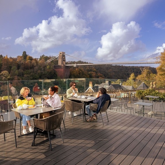 Friends enjoying tea and cake on a rooftop terrace, overlooking a river and suspension bridge.
