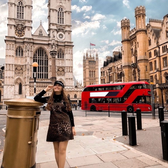 A girl standing by a golden letter box with a red bus behind