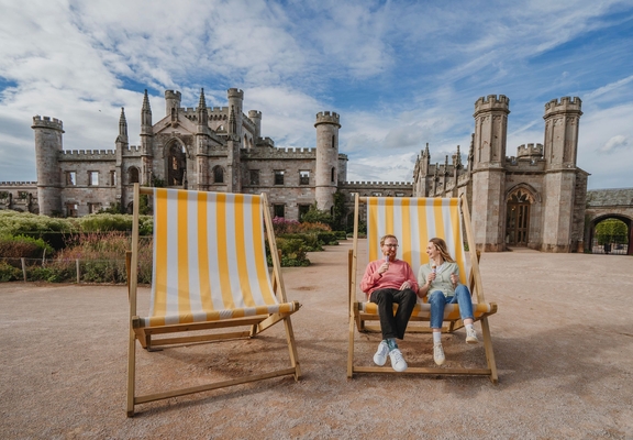 Man and woman holding ice creams in oversized deck chairs in manicured castle grounds and gardens.