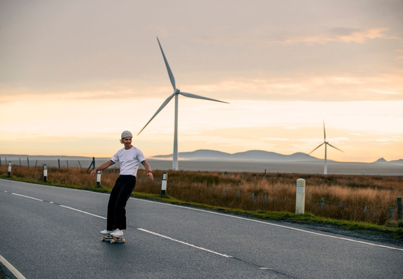Un uomo con lo skateboard si gode la superficie stradale piatta su cui pattinare nelle Highlands scozzesi, con le turbine eoliche alle spalle
