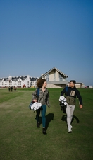 A man and a woman carrying golf equipment walking on the grounds of a country golf club.