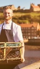 Chef wearing apron on pier holding lobster trap with lobster