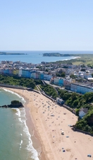An overhead shot of a sandy beach lined with sunbathers, surrounded by brightly coloured townhouses and countryside.