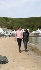 Two people walking along a sandy beach with the ocean to one side and pubs and shops on the other side. 