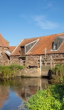 A stone water mill with an unusual conical roof beside a pool of water.