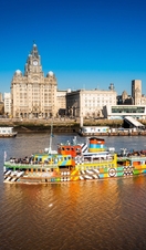 A colourful ferry crossing a river with a city backdrop and bright blue sky