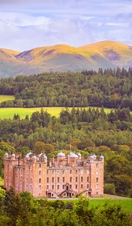 Exterior of a sandstone palace with ornate turrets surrounded by woodland with high mountains in the distance in autumn.