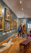 A man and a woman stand looking at paintings in a gallery