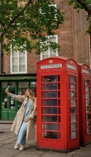 A woman leans against a red telephone box and takes a selfie in a town square.
