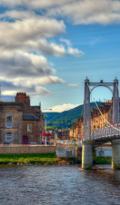 A suspension bridge with people walking across spanning a wide river with houses on the other side