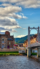 A suspension bridge with people walking across spanning a wide river with houses on the other side