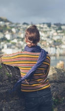 A young woman is standing by a stone wall in a village and is looking at the boats moored in the estuary