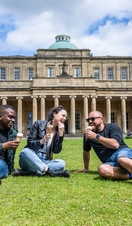 Two men and a woman sit on grass outside of a heritage building