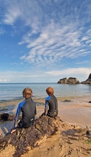 Back view of two young boys in wetsuits sitting on a rock looking out to sea with blue sky