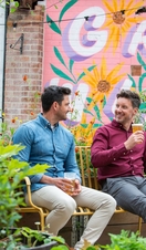 Two men sat on a bench in a garden with a drink