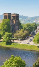 A cathedral with two towers overlooks a river on a sunny day, with trees in the foreground.