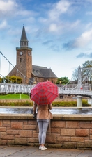 A woman with an umbrella facing a river next to a bridge towards a church