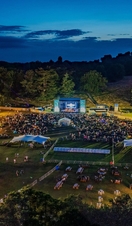 A large outdoor concert with a large screen with crowds and many tents and fairground rides.