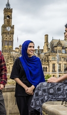 Two women and a man stand and talk in front of a town view