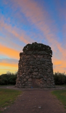 A small stone tower with a plaque on it surrounded by grass and moor under a vibrant blue and orange sky at sunset.
