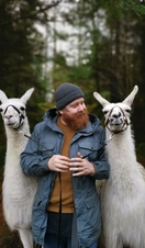 A bearded man wearing a wool hat holding two white alpacas