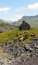 A slate house in the middle of the Snowdonia Slate Trail surrounded by green mountains, with blue sky and clouds