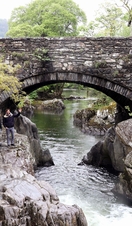 Two men waving from some rugged rocks beneath a stone bridge over a narrow river