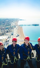 Group of friends at the top of i360 pod in climbing gear with coastline of Brighton. below