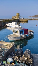 Two small boats moored in a small stone harbour on a sunny day, with seven lobster cages in the foreground.