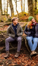 A man and woman in hiking gear smile at each other while sitting on tree roots in a forest.