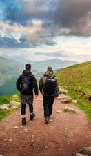 Two male hikers trekking along a pebbled path, cloudy skies, green rolling mountain vista.