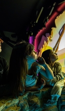 A man and two young girls watch an animated water monster with a long neck on a screen .