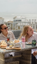 Two men and two women laugh together while sat on a balcony eating lunch with the sea beyond