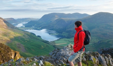 A man wearing a rucksack stands on a mountain fell overlooking hills and a lake