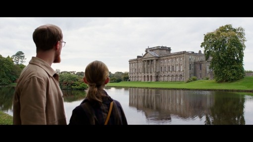 A man and a woman stand across a lake looking toward a heritage house