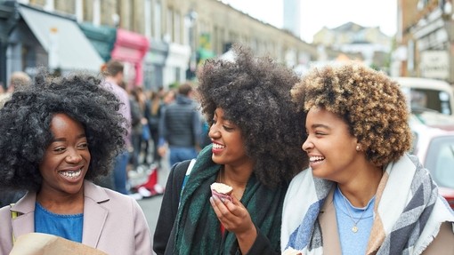 Three young mixed race female friends laughing and having fun in a city street eating cupcakes, Columbia Road, London