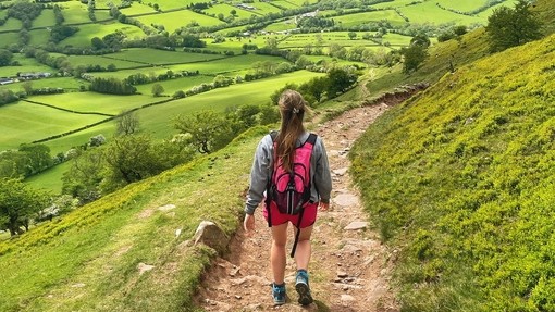 Back view of young woman walking on a path through a lush green valley and hills